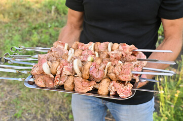 Pieces of raw pork strung on skewers lie on an iron tray held by a man, ready for grilling or...
