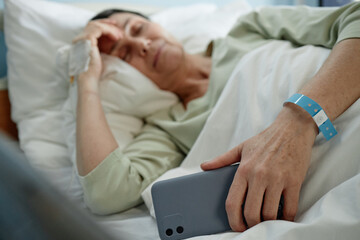 Elderly woman reclining in hospital bed holding smartphone in hand, wearing wristband and resting head on pillow. Appears visibly tired and in recovery state