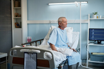 Fototapeta premium Elderly male patient sitting on hospital bed while undergoing medical treatment in a brightly lit room setting with medical equipment around him