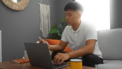 Young chinese man working indoors on a laptop while holding a smartphone in a cozy living room setting.