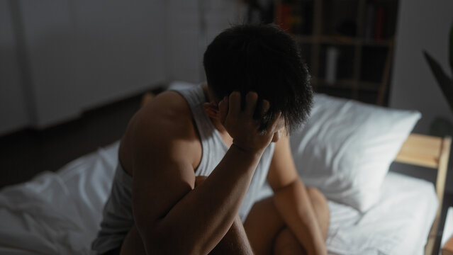 Young man in a bedroom sits on a bed with his head in his hand, appearing thoughtful or stressed, illuminated by soft indoor lighting.