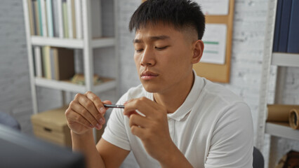 Young, handsome chinese man sitting in his bedroom, focusing intently on a pen in his hands, surrounded by bookshelves and personal items, creating a thoughtful and introspective atmosphere. © Krakenimages.com
