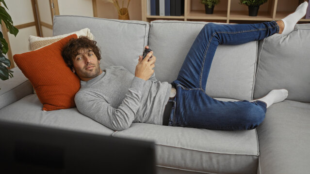 Handsome man relaxing on a couch with a gaming controller in a modern living room setting, demonstrating a laid-back and stylish vibe at home.