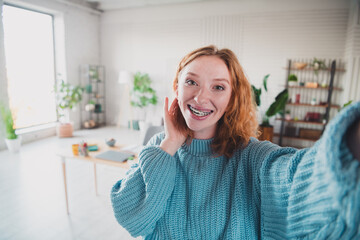 Smiling young woman with red hair takes a selfie in a cozy, sunlit living room wearing a casual knitted pullover.