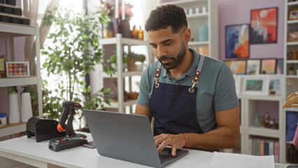 Young man in apron working on laptop in home decor store surrounded by plants and artwork