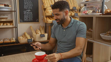 Young man sitting in a bakery, drinking coffee and using phone, with bread in the background
