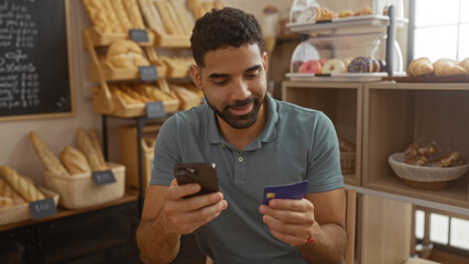 Young hispanic man using phone and credit card in a bakery surrounded by various breads and pastries