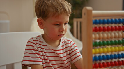 Blond boy playing with abacus indoors in kindergarten looking focused wearing striped clothing