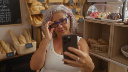 Woman taking selfie in bakery with variety of breads and pastries in background, wearing glasses, grey haired, elderly, beautiful, mature, hispanic, indoors, and smiling.