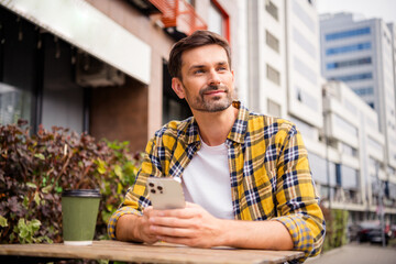 Young casual man in yellow checkered shirt enjoying city outdoor lifestyle while holding smartphone and coffee at a cafe on a sunny day.