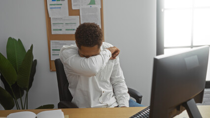 Young man sneezing into elbow in office setting with computer on desk and bulletin board in background