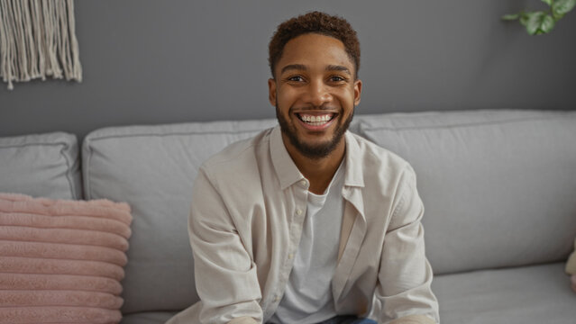 Young african american man sitting in living room smiling happily at home interior sitting on grey sofa. - Powered by Adobe
