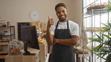 Handsome young man wearing an apron smiling and giving a thumbs-up in a cozy bakery with shelves of pastries and cakes