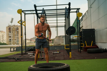Muscular athlete using sledgehammer to hit tire during intense outdoor workout session,...