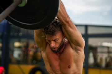 Obraz na płótnie Canvas Muscular male athlete lifts a weight plate outdoors, stretching his back and biceps while engaging in a challenging workout to enhance his physical fitness
