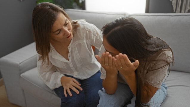 Mother comforting daughter in living room with a loving gesture, indoors at home, portraying family support and emotional bond between two women.