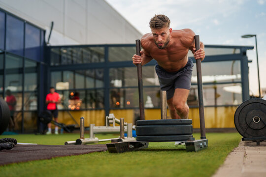 Muscular sportsman performing intense physical exercise with weighted sled on a green turf field in an outdoor gym setting, demonstrating strength and dedication to fitness