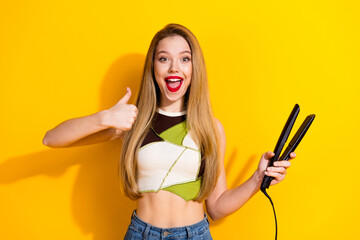 Cheerful young woman with long straight hair giving thumbs-up while holding hair straightener...