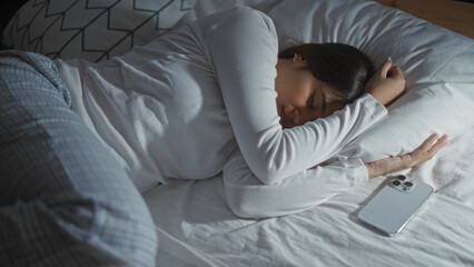 Woman sleeping peacefully in a home bedroom on a bed next to a smartphone during the night