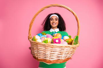 Young woman holding basket with colorful eggs in cheerful pink background wearing green sweater and...