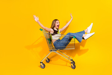 Cheerful young woman enjoying a fun moment riding in a shopping cart against a vibrant yellow...