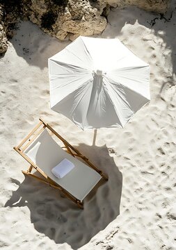 Fototapeta A high-angle shot of a white beach umbrella and chair on sandy shores, showcasing minimalist design with sunlight and shadows in neutral tones. Perfect for summer and relaxation themes.