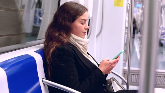 Cheerful young woman in a black coat and beige scarf sitting inside a modern subway train, laughing while looking at her smartphone. She enjoys a fun moment during her commute. 