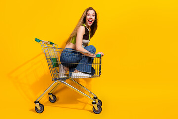 Young woman sitting in a shopping cart against a vibrant yellow background, expressing joyful and...