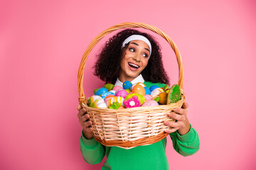 Young cheerful woman in green jumper holding colorful Easter egg basket with pink backdrop