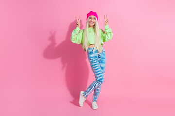 Young female in vibrant casual fashion posing against a pink background displaying peace gestures with a cheerful and stylish expression