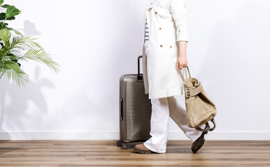 Beige plastic suitcase on wheels in woman hands in white trench coat and trousers and casual backpack, ready for trip or travel