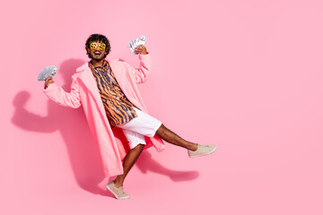 Young stylish man in trendy pink coat showing fan of cash on pink background demonstrating joyful expression and modern casual fashion
