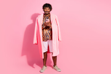 Young stylish man with curly hair in a printed shirt and pink coat using a smartphone against a bright pink background