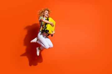 Young woman jumping joyfully against an orange background holding a yellow suitcase, expressing happiness and energy