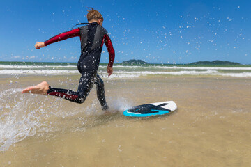 Young surfer in a wetsuit running to skimboard on the shoreline, splashing through shallow water....