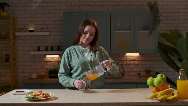 Woman at the table in the kitchen preparing healthy vegetable salad for dinner, pouring fresh orange juice from squeezer in a glass and drinks it. Slow motion