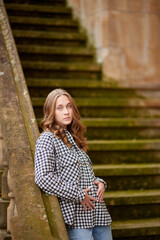 Stylish young woman posing on an old mossy staircase in a European town with red window shutters, wearing a checkered flannel shirt and blue jeans.