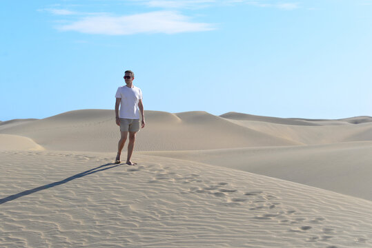 A young man walking along a sand dune through barkhan in Maspalomas, Gran Canaria on a hot sunny day. Desert travel touristic concept