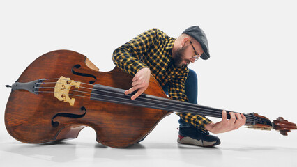 Dynamic musician with hat and glasses performing on double bass, crouching low while plucking strings with energy. Concept of music, jazz, concert, festival, talent, performance.
