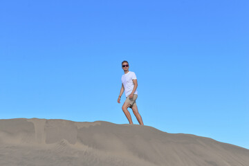 A young man walking along a sand dune through barkhan in Maspalomas, Gran Canaria on a hot sunny day. A self confident man goes up the mountain - success and achievements concept