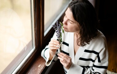 Woman relaxing indoors, enjoying the scent of lavender bouquet by the window during a peaceful afternoon