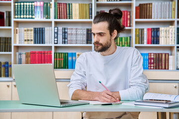 Young male teacher working in library sitting at desk with laptop books
