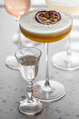 A cocktail with a passion fruit slice garnish, served with a small glass of clear liquid on a light surface, blurred drinks in the background, perfect for bar and mixology themes.