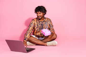 A young man sitting against a pink background holding a gift box and interacting with a laptop,...