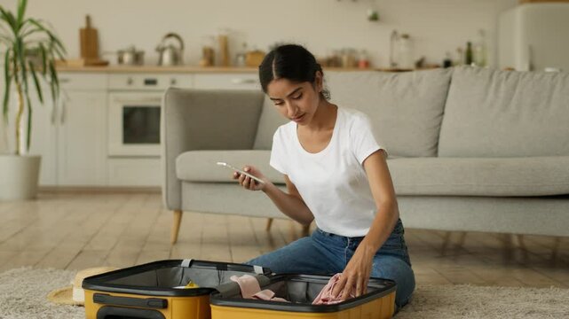 Indian woman Arabian lady female girl at home preparing to trip vacation travel checking items of things clothes packing cloth using mobile phone online list remember put summer hat inside traveling