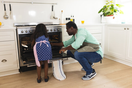 African American father and daughter baking together in kitchen, checking oven with excitement