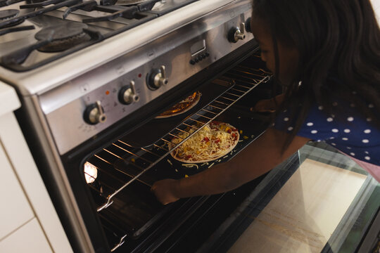 Young girl carefully placing homemade pizza in oven, learning cooking skills at home - Powered by Adobe