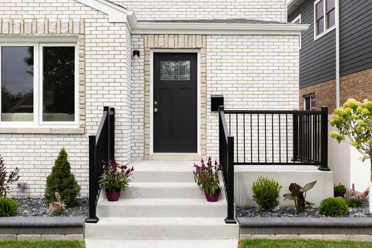 A front door detail on a white brick home with concrete steps and black railing leading to a black front door with a mosaic window.