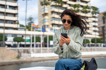 Fototapeta premium A fashionable young woman with sunglasses and a quilted jacket sits near the waterfront, smiling as she types on her smartphone, with flags and modern buildings in the background