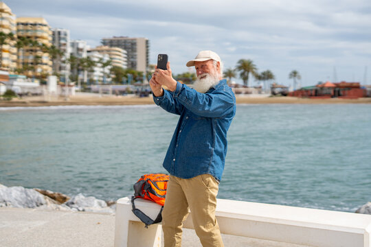 An elderly man with a white beard, wearing a beige cap and denim shirt, takes a selfie with his smartphone by the waterfront, with a Spanish flag, palm trees, and a coastal town in the background